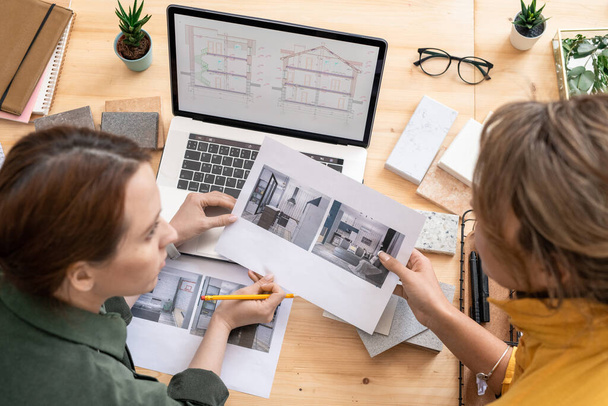 Zwei junge kreative Unternehmerinnen sitzen am Holztisch und diskutieren Fotos aus dem heimischen Interieur, während sie an einer neuen Ordnung arbeiten - Foto, Bild