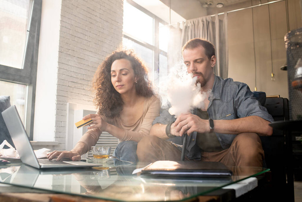 Jovem contemporâneo fumando cigarro eletrônico enquanto se senta ao lado de sua esposa fazendo compras on-line na frente de laptop e cartão de plástico usina
 - Foto, Imagem