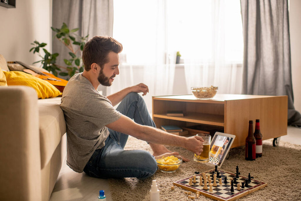 Позитивний бородатий чоловік сидить на підлозі і п'є пиво з другом, спілкуючись онлайн в домашній ізоляції
 - Фото, зображення