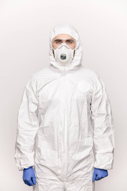 Contemporary young man in protective eyeglasses, blue gloves, white coveralls and respirator standing in front of camera in isolation - 写真・画像