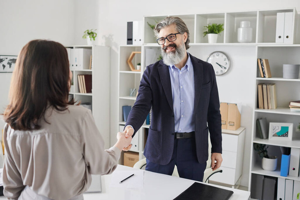 Happy and successful professional in formalwear and eyeglasses shaking hand of one of his clients over desk after making an agreement - Photo, Image