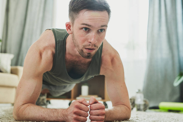 Młody mężczyzna w odzieży sportowej stojący na desce podczas ćwiczeń na podłodze salonu w okresie kwarantanny lub samoizolacji - Zdjęcie, obraz