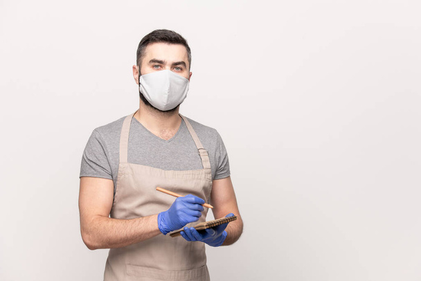 Young seious waiter in apron, protective mask and gloves writing down your order in notepad while working in cafe or restaurant - Photo, Image