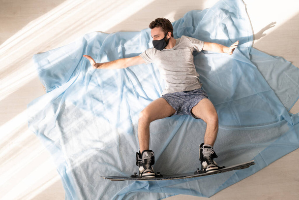 Jonge snowboarder in gezichtsmasker op de vloer thuis en beeldvorming dat hij snowboardt in de bergen na quarantaine - Foto, afbeelding