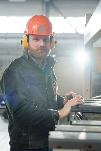 Portrait d'un homme brutal en vêtements de travail et protecteurs d'oreilles travaillant avec des pièces métalliques dans un atelier d'usine - Photo, image