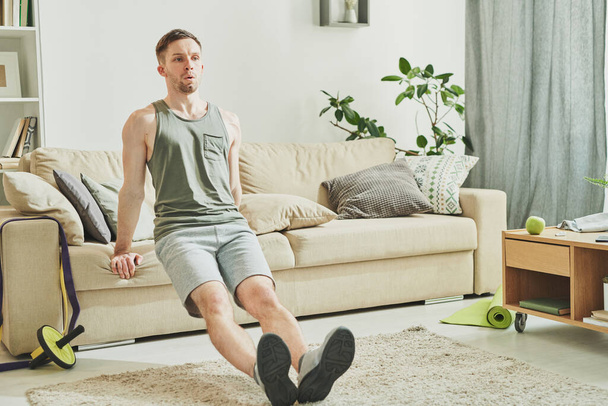 Joven hombre activo en ropa deportiva sosteniendo por sofá a sus espaldas mientras hace ejercicio difícil por la mañana durante el período de estancia en casa
 - Foto, Imagen