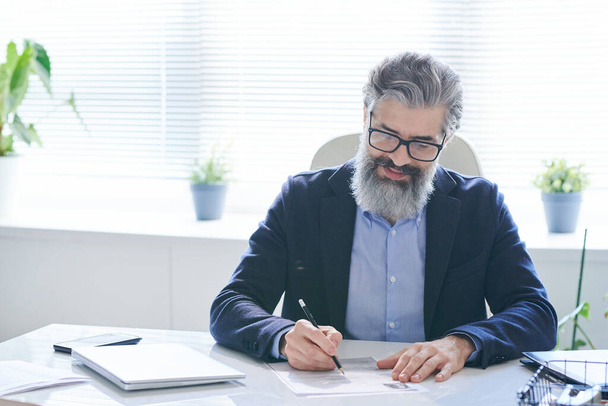 Doświadczony profesjonalista w okularach i odzieży formalnej robi notatki na papierze siedząc przy biurku przy oknie i czekając na klientów - Zdjęcie, obraz
