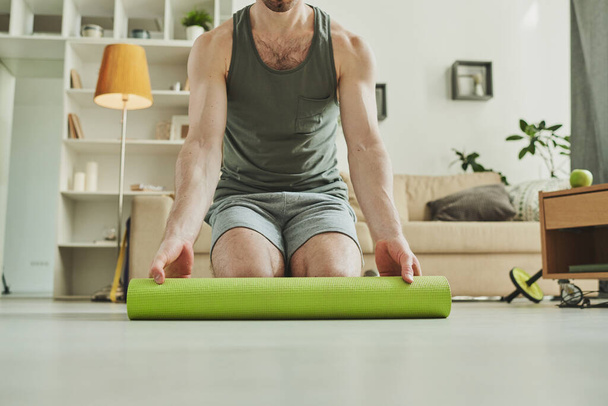 Νέοι σύγχρονος αθλητής στέκεται στα γόνατά του, ενώ ξετυλίγεται πράσινο χαλί για την άσκηση στο πάτωμα στο περιβάλλον στο σπίτι - Φωτογραφία, εικόνα