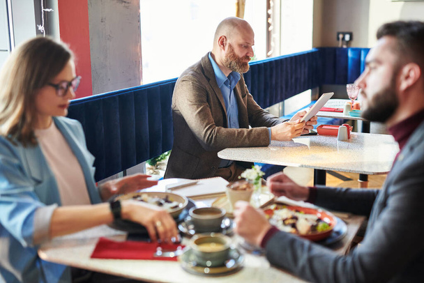 Серьезный лысый человек сидит за столом и с помощью планшета подключен к Wi-Fi во время работы в современном кафе
 - Фото, изображение