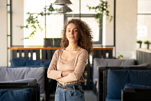 Young successful businesswoman with her arms crossed by chest standing inside luxurious cafe or restaurant with couches on both sides - Foto, Imagen