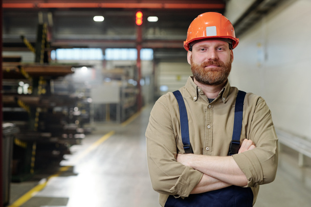 Portrait of content brutal bearded factory man in orange hardhat standing with crossed arms in workshop - Foto, afbeelding
