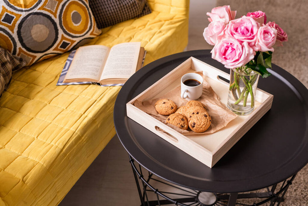 Домашнє натюрморт з купою рожевих троянд, домашньою закускою та напоєм на маленькому столі та відкритою книгою та подушками на жовтому дивані
 - Фото, зображення