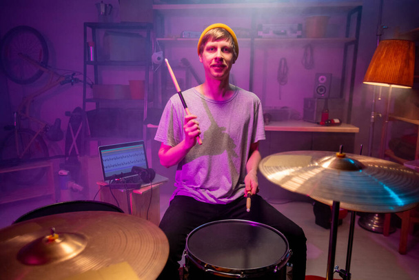 Young man in tee, jeans and beanie looking at you while hitting drums and cymbals with drumsticks and recording his music on laptop - Fotó, kép