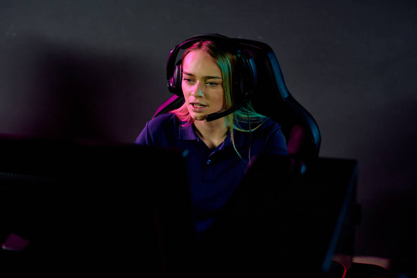 Jovem loira séria fêmea e-sport gamer no fone de ouvido olhando para a tela do computador enquanto sentado no clube escuro na frente do monitor
 - Foto, Imagem