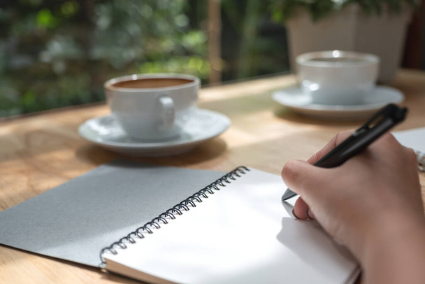 Closeup image of a hand writing down on a white blank notebook with coffee cup on wooden table  - Foto, Bild