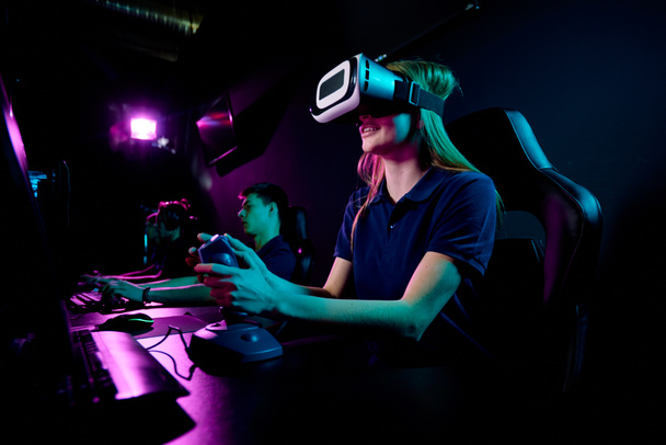 Contemporary eenage girl in vr headset using joystick while sitting in front of computer monitor and playing new video game in club - Foto, Imagen