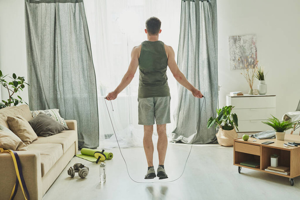 Vista trasera del joven en ropa deportiva saltando con una cuerda mientras está parado frente a la ventana de la sala de estar durante el entrenamiento
 - Foto, Imagen