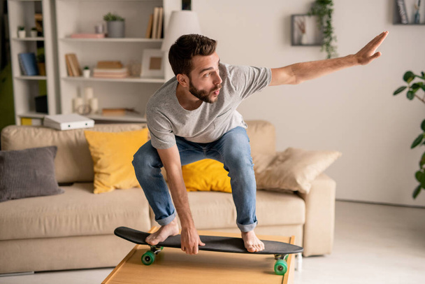Екстатичний молодий бородатий чоловік стоїть на скейтборді, балансуючи на кавовому столі, розважаючись у карантині
 - Фото, зображення