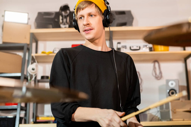 Jovem baterista loiro contemporâneo em fones de ouvido, gorro amarelo e camisola preta batendo pratos e tambor na garagem
 - Foto, Imagem