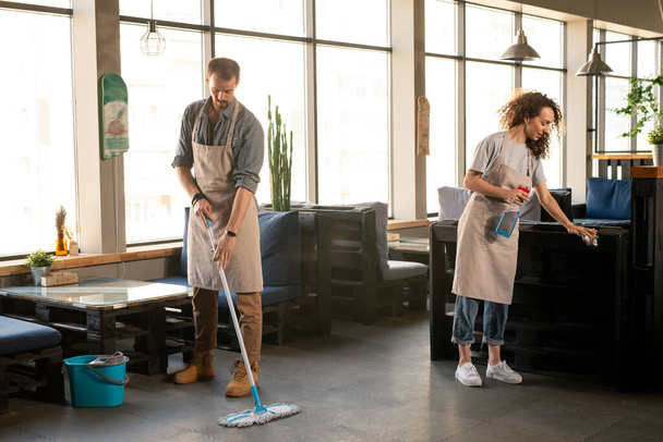 Zwei Besitzer eines modernen Restaurants oder Cafés waschen den Boden und desinfizieren Möbel nach dem Arbeitstag mit Desinfektionsmittel - Foto, Bild