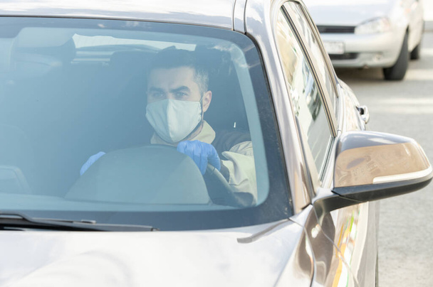 Young serious courier in protective mask and gloves sitting in his car and driving to clients while delivering goods during quarantine - Fotografie, Obrázek