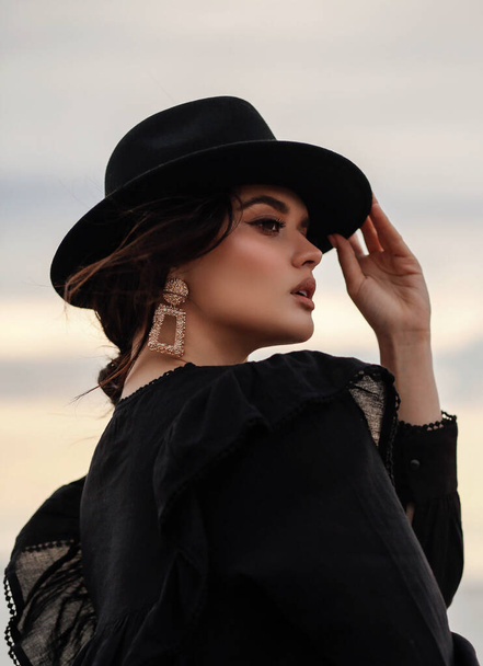 fashion outdoor photo of beautiful woman with dark hair in elegant clothes and black hat posing on the beach - Foto, Imagem