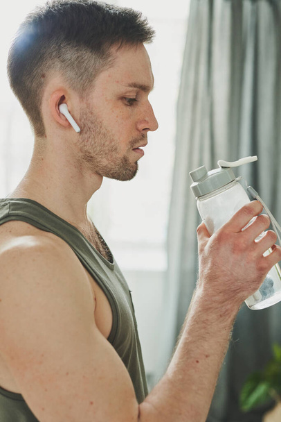 Fiatal férfi fülhallgatóban és sportmellényben, aki egy üveg vizet tart a szája mellett, miközben megissza, miután keményen edzett otthon. - Fotó, kép