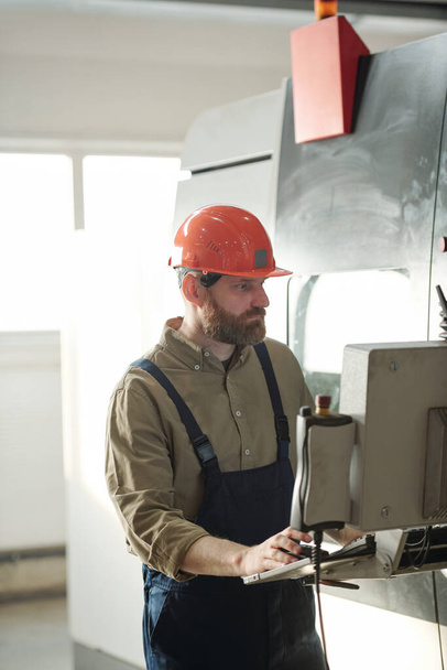 Concentrated bearded operator in hardhat and overalls choosing setups on CNC machine computer while working in factory shop - Photo, Image