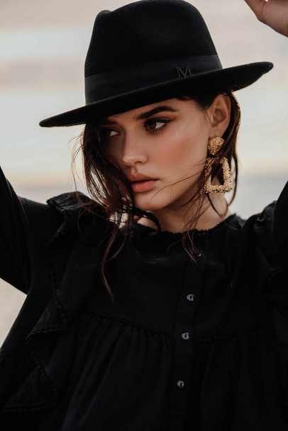 fashion outdoor photo of beautiful woman with dark hair in elegant clothes and black hat posing on the beach - Fotó, kép