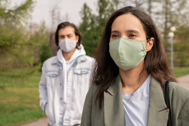 mooie jonge brunette vrouw in beschermende masker wandelen in openbaar park tijdens pandemie periode op de achtergrond van casual man - Foto, afbeelding