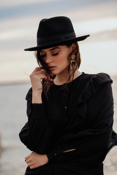 fashion outdoor photo of beautiful woman with dark hair in elegant clothes and black hat posing on the beach - Zdjęcie, obraz