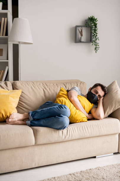Joven triste en máscara de tela acostado con almohada amarilla en el sofá en aislamiento coronavirus
 - Foto, Imagen