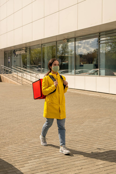 Contemporary courier in casualwear and protective mask moving along modern building on sunny day while delivering orders to clients - Fotografie, Obrázek