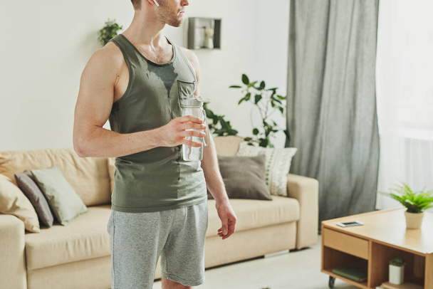Молодий м'язистий чоловік у спортивному одязі тримає пляшку води на грудях, маючи перерву між наборами тренувань в домашньому середовищі
 - Фото, зображення