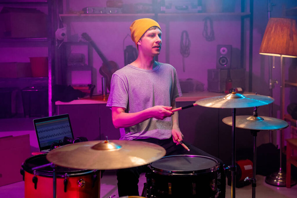 Hedendaagse jonge muzikant in casualwear slaan bekkens en drums met drumstokken tijdens het opnemen van zijn muziek in de garage of studio - Foto, afbeelding