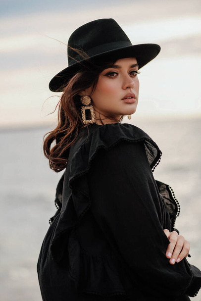 fashion outdoor photo of beautiful woman with dark hair in elegant clothes and black hat posing on the beach - Photo, Image