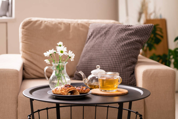 Bunch of white flowers in jug with water, snack on plate and green herbal tea in cup and teapot on small table by armchair with pillow - Foto, Bild