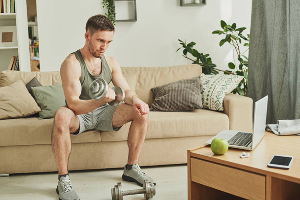 Молодой человек в спортивной форме сидит на диване перед ноутбуком, делая упражнения с гантели для бицепсов в гостиной
 - Фото, изображение