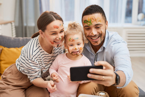 Молодой человек и женщина делают смешное селфи на смартфоне со своей маленькой дочерью с гуашью на лицах
 - Фото, изображение