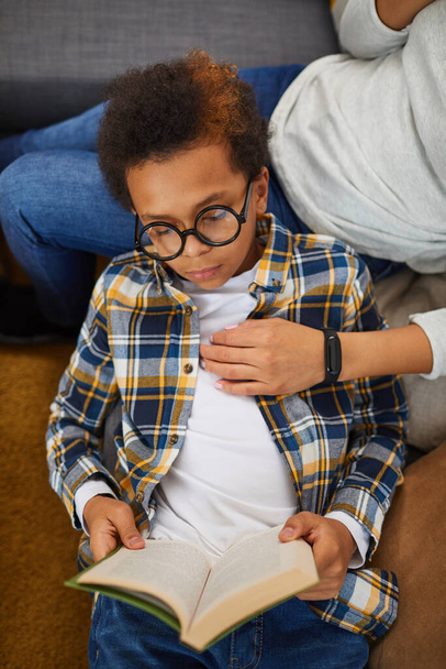 Top view ritratto di carino ragazzo africano indossa grandi occhiali libro di lettura mentre sdraiato sul divano con la mamma, concetto di homeschooling - Foto, immagini