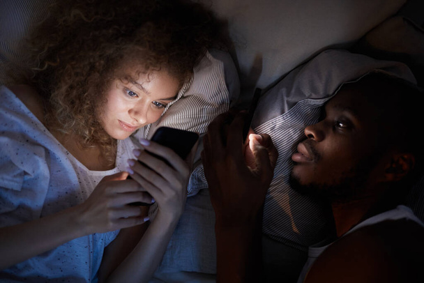 Πάνω άποψη στο νεαρό ζευγάρι μικτή φυλή χρησιμοποιώντας smartphones, ενώ βρίσκεται στο κρεβάτι τη νύχτα, επικεντρωθεί στην χαμογελαστή σγουρά μαλλιά γυναίκα - Φωτογραφία, εικόνα