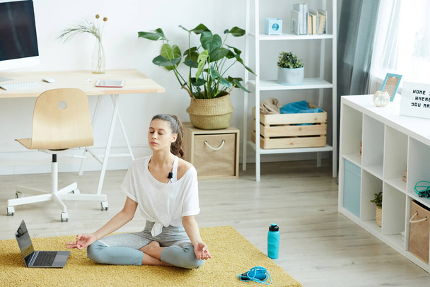 Täyspitkä muotokuva nykyajan nuori nainen meditoi kotona istuessaan lootus asennossa aikana jooga harjoitus, kopioi tilaa - Valokuva, kuva