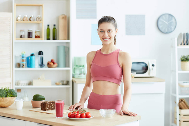 Spor yapan genç bir kadının belden yukarı portresi mutfağın içinde sağlıklı yemekler pişiriyor ve kameraya gülümsüyor. - Fotoğraf, Görsel