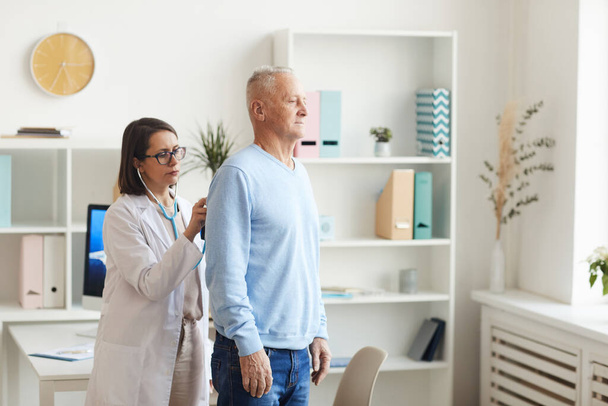 Seitenansicht Porträt einer Ärztin, die dem Herzen und der Atmung eines älteren Patienten zuhört, Stethoskop während der Konsultation in der Klinik aufsetzt, Kopierraum - Foto, Bild