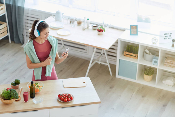Vue grand angle à la jeune femme contemporaine en utilisant un smartphone tout en cuisinant des aliments de fitness dans l'intérieur de la cuisine, copier l'espace - Photo, image