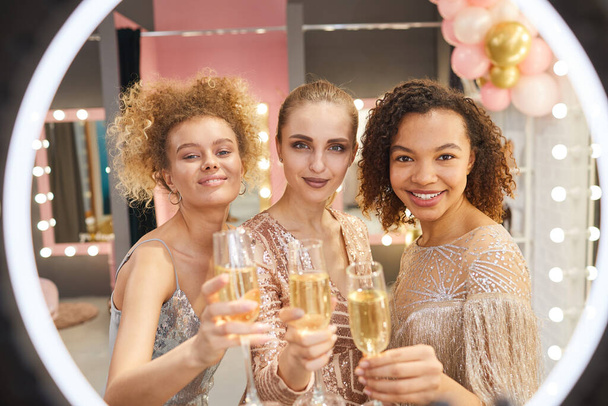 Gyönyörű fiatal nők soknemzetiségű csoportja pezsgős poharakkal és mosollyal az öltözőben, miközben a kamerát nézik, gyűrűs fényben. - Fotó, kép
