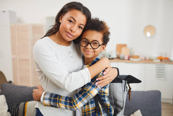 Cintura até retrato de feliz mãe afro-americana abraçando filho ao enviá-lo para a escola em setembro, ambos olhando para a câmera
 - Foto, Imagem