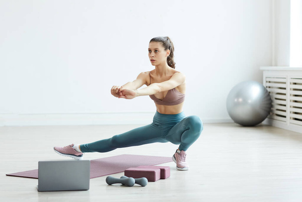 Minimale volledige lengte portret van pasvorm jonge vrouw stretching benen terwijl u geniet van sport workout thuis in witte kamer, kopiëren ruimte - Foto, afbeelding