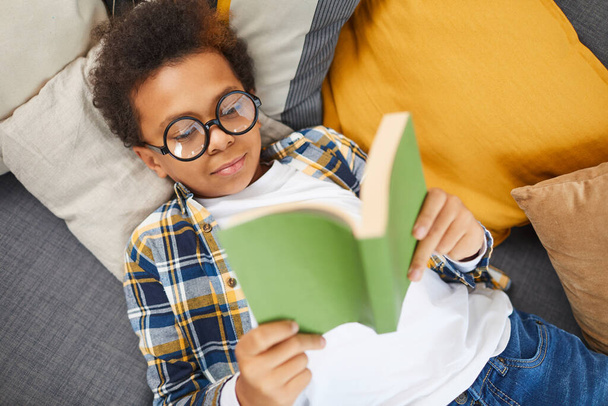 Над просмотром портрет симпатичного африканского мальчика в больших очках, читающий книгу, лежа на диване, домашнее обучение концепции, копировать пространство
 - Фото, изображение