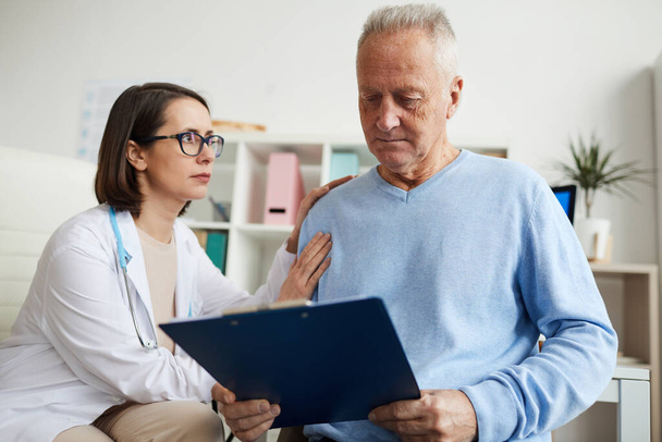 Portrait of caring female doctor comforting senior patient reading diagnosis on clipboard during consultation in clinic, copy space - Photo, Image